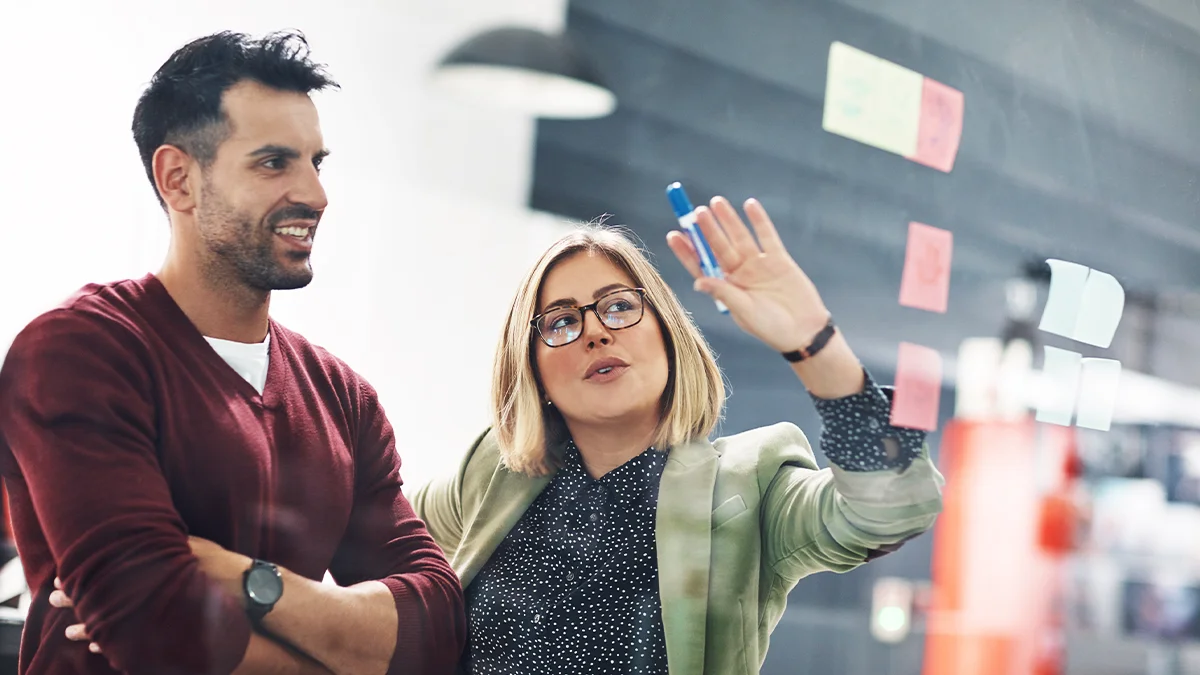 Two marketers brainstorming using sticky notes on glass wall.